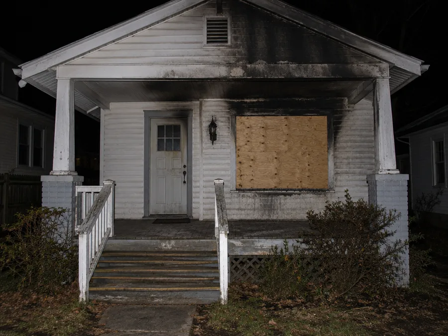 The front porch of an older wood-sided bungalow at night, plywood screwed over the front window and black soot streaking up the siding above it