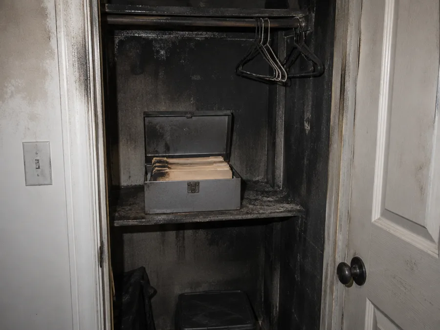 A smoke-blackened hallway closet with its door open and a grey metal document box on the shelf, holding a stack of folders with sooty edges