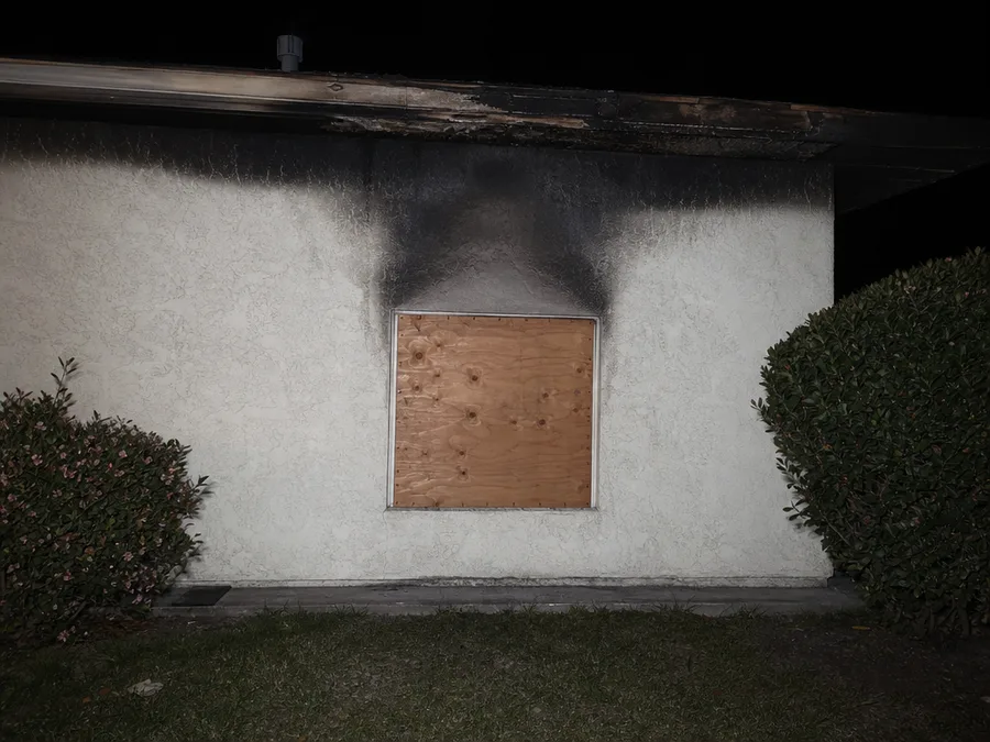 A window opening covered with screwed plywood, soot streaking the stucco above it